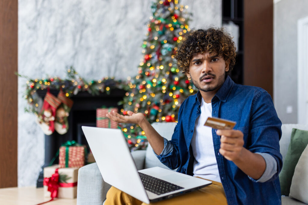 Portrait of cheated unsatisfied man at home on New Year and Christmas holidays, hispanic man looking at camera with laptop rejected money transfer, using bank credit card.